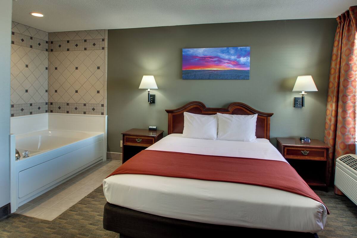 A hotel room with a large bed, a bathtub, two bedside tables with lamps, and a painting on the wall.
