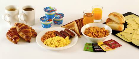 A breakfast spread with croissants, sausages, scrambled eggs, toast, yogurt, juice, and cheese.
