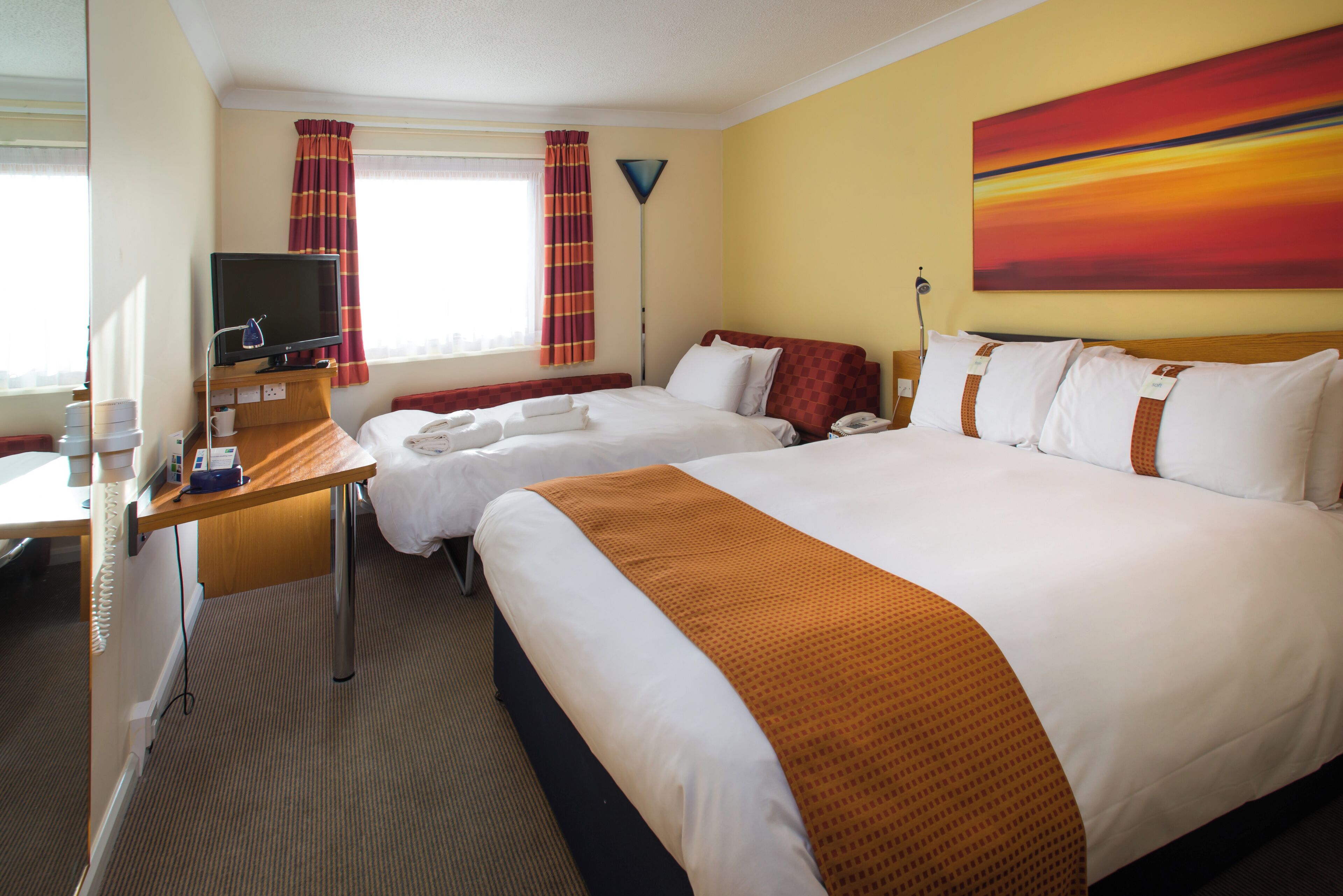 A hotel room with two beds, a desk with a computer, a TV, and a painting on the wall.