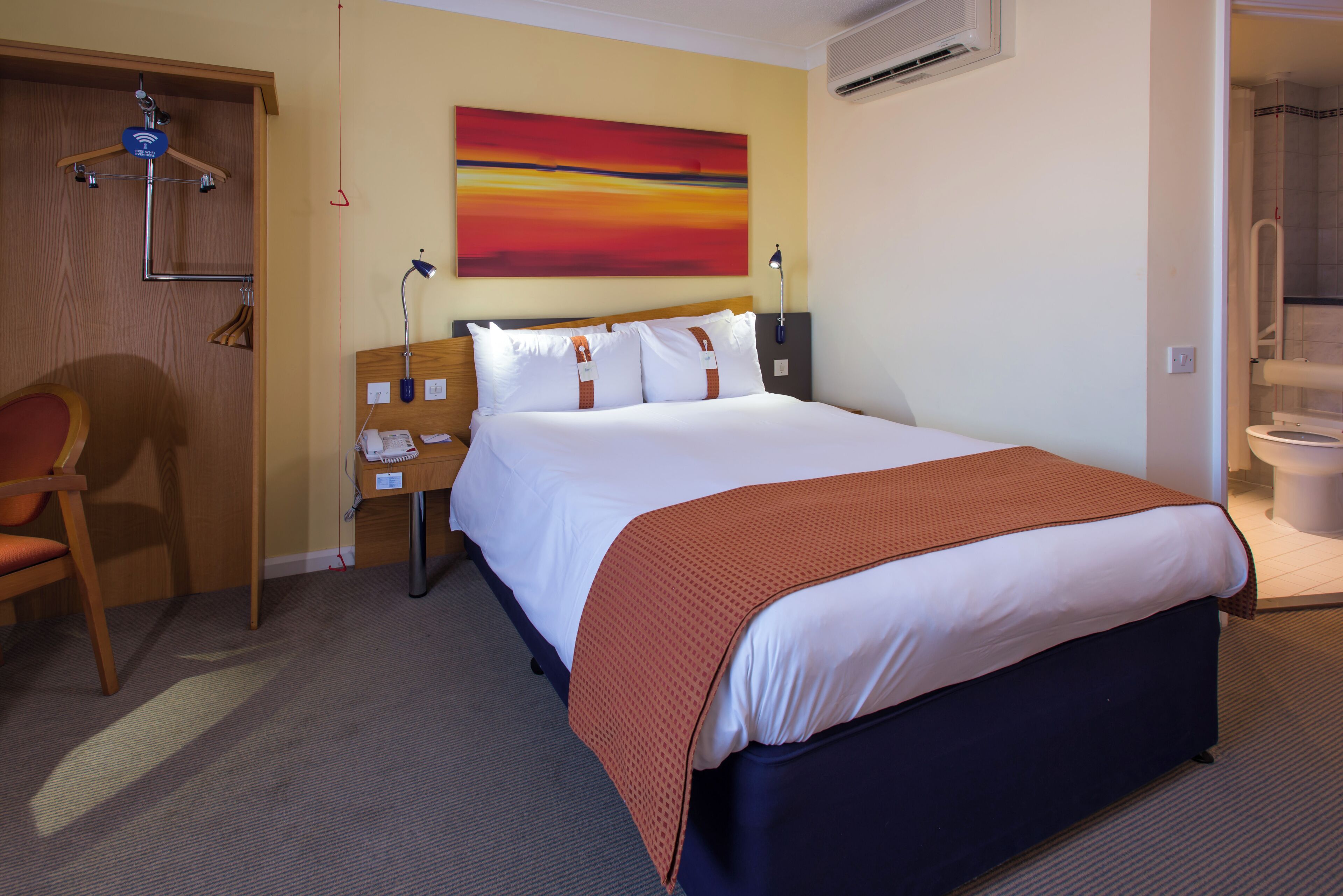 A hotel room with a bed, a chair, a desk, a wall-mounted air conditioner, and a painting.