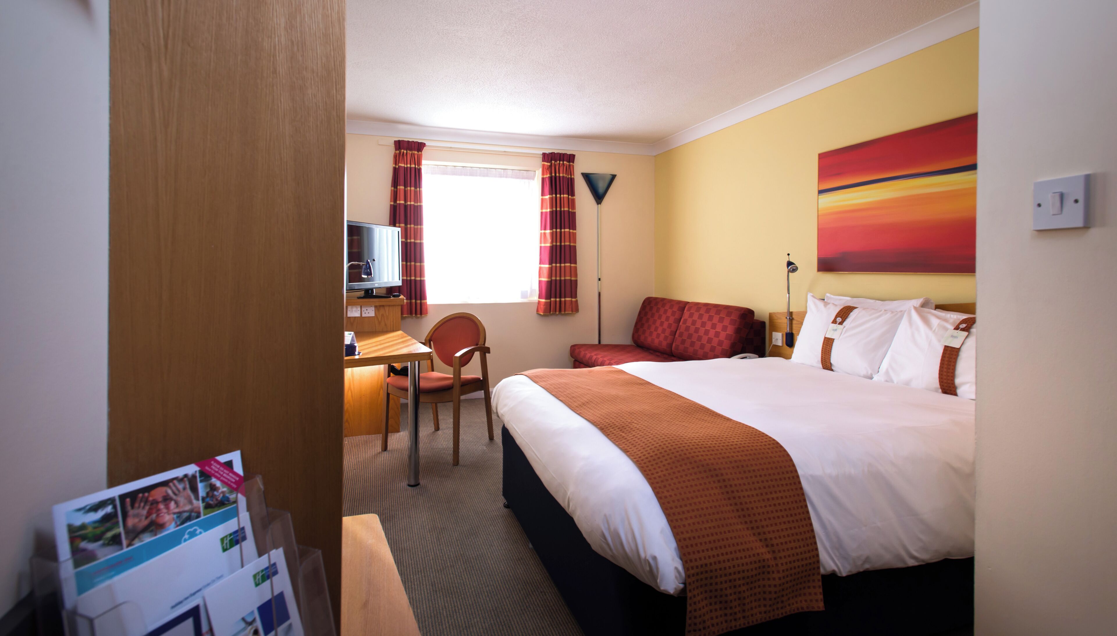 A hotel room with a bed, a desk, a chair, a television, and a painting on the wall.