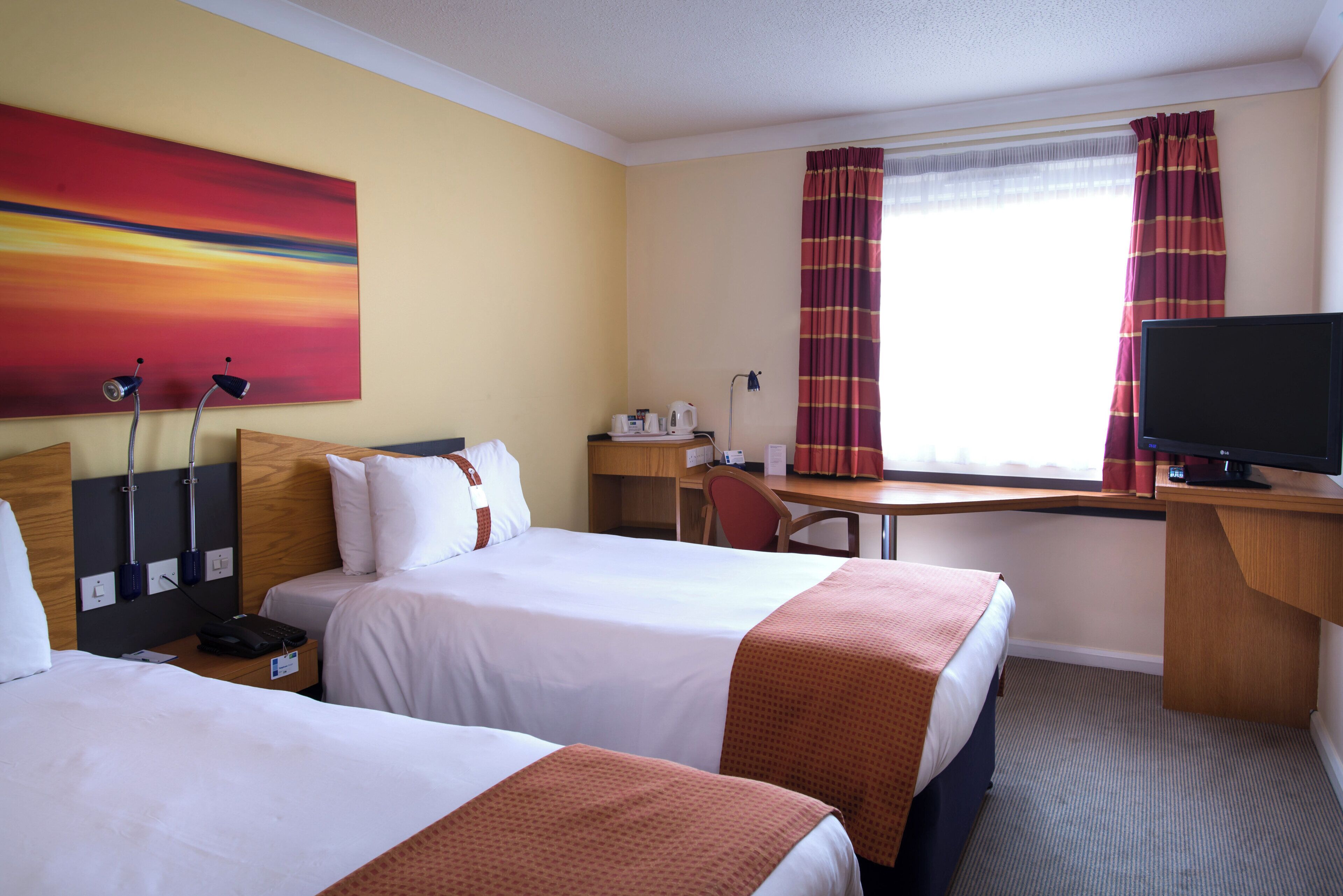A hotel room with two beds, a desk, a television, and a painting on the wall.