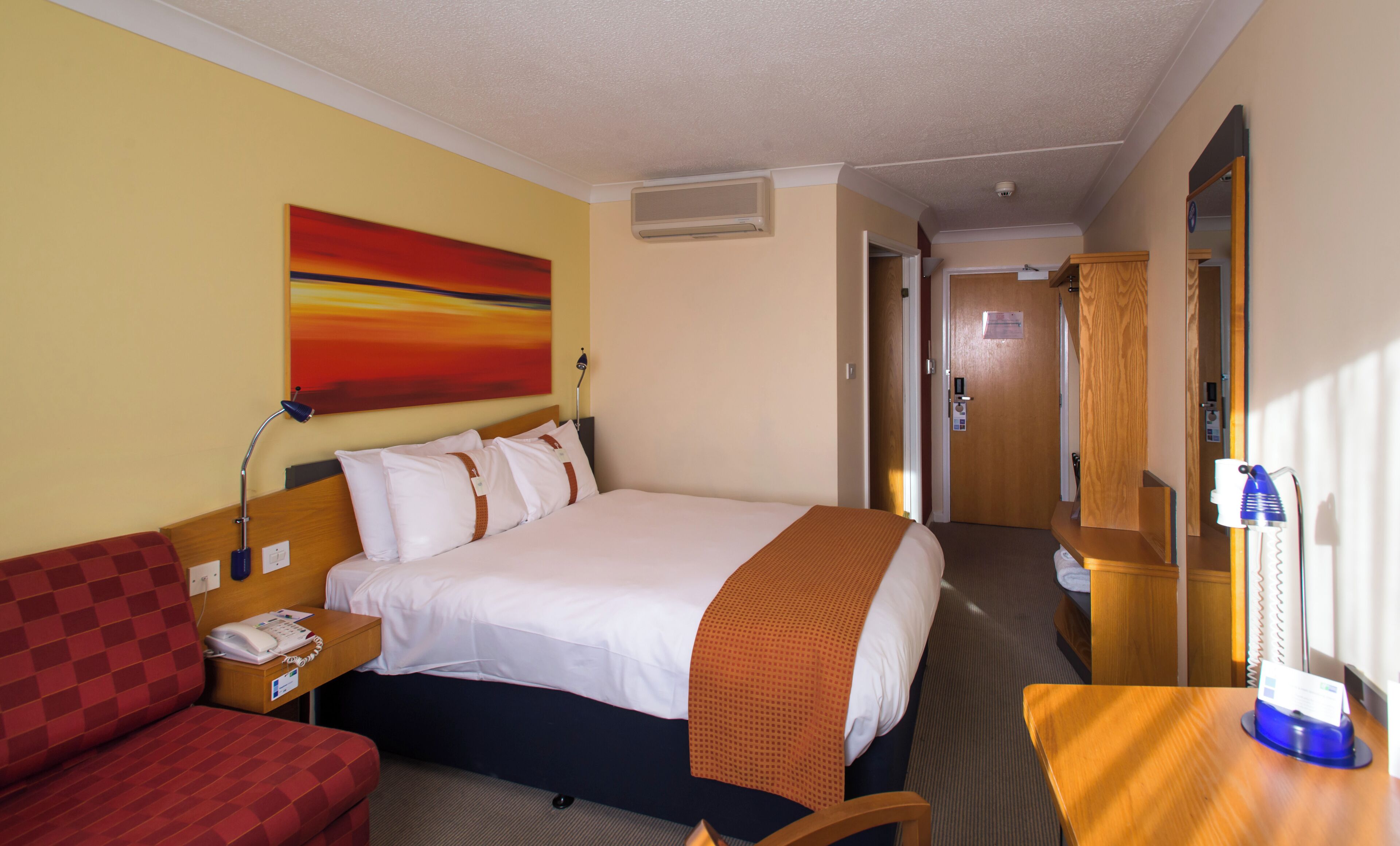 A hotel room with a bed, a desk, a telephone, a mirror, and a door leading to another room.