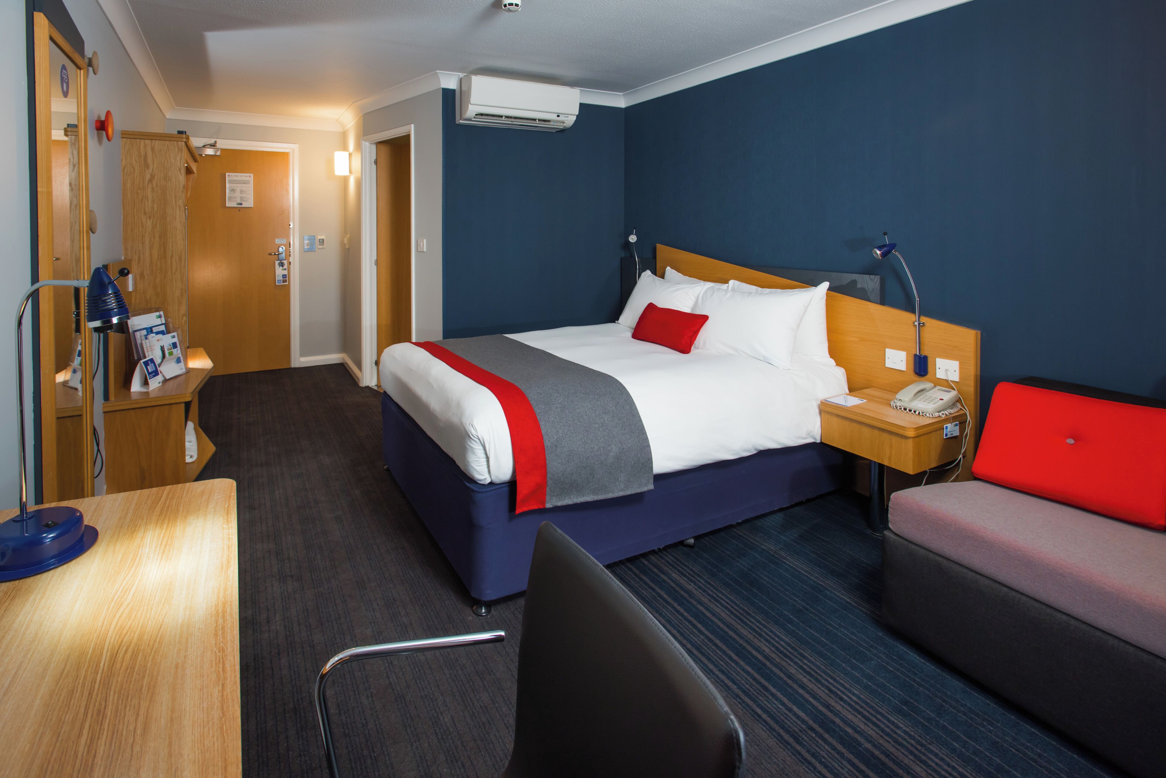 Habitación de hotel con cama, escritorio, silla, mesita de noche con lámpara y un sofá rojo.