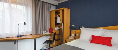 Una habitación de hotel con cama, escritorio, silla, armario y ventana con cortinas.