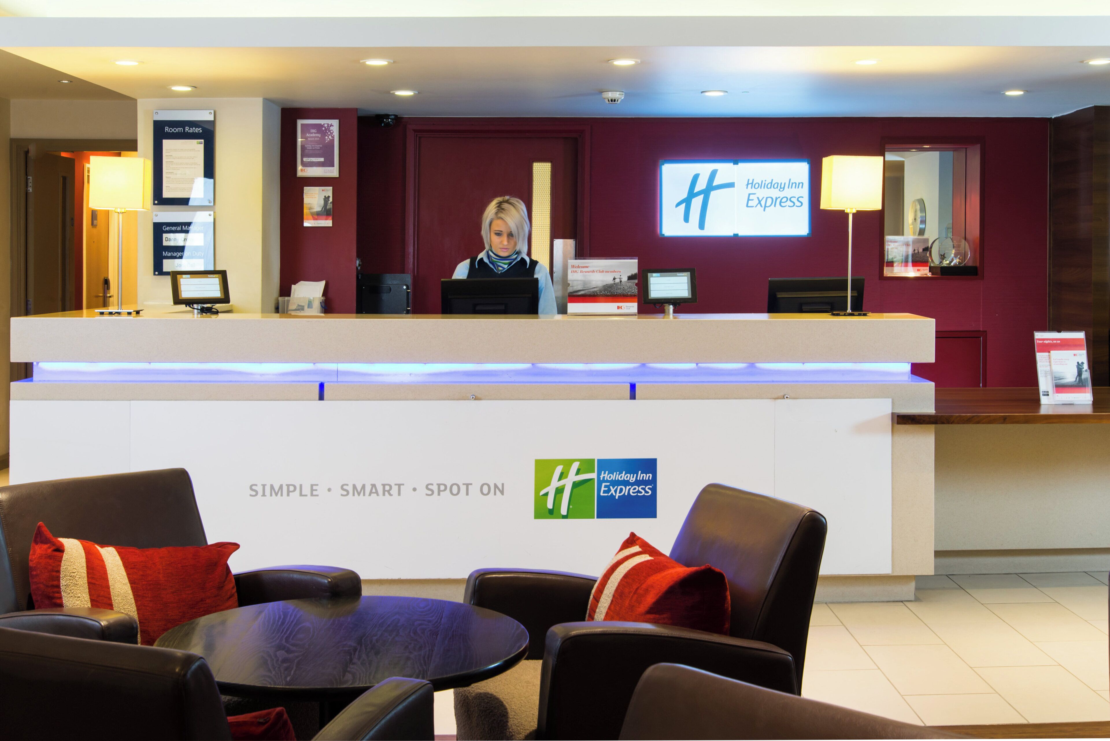 Mostrador de recepción de Holiday Inn Express, con recepcionista, pantalla digital y zona de estar con sillas y una mesa.