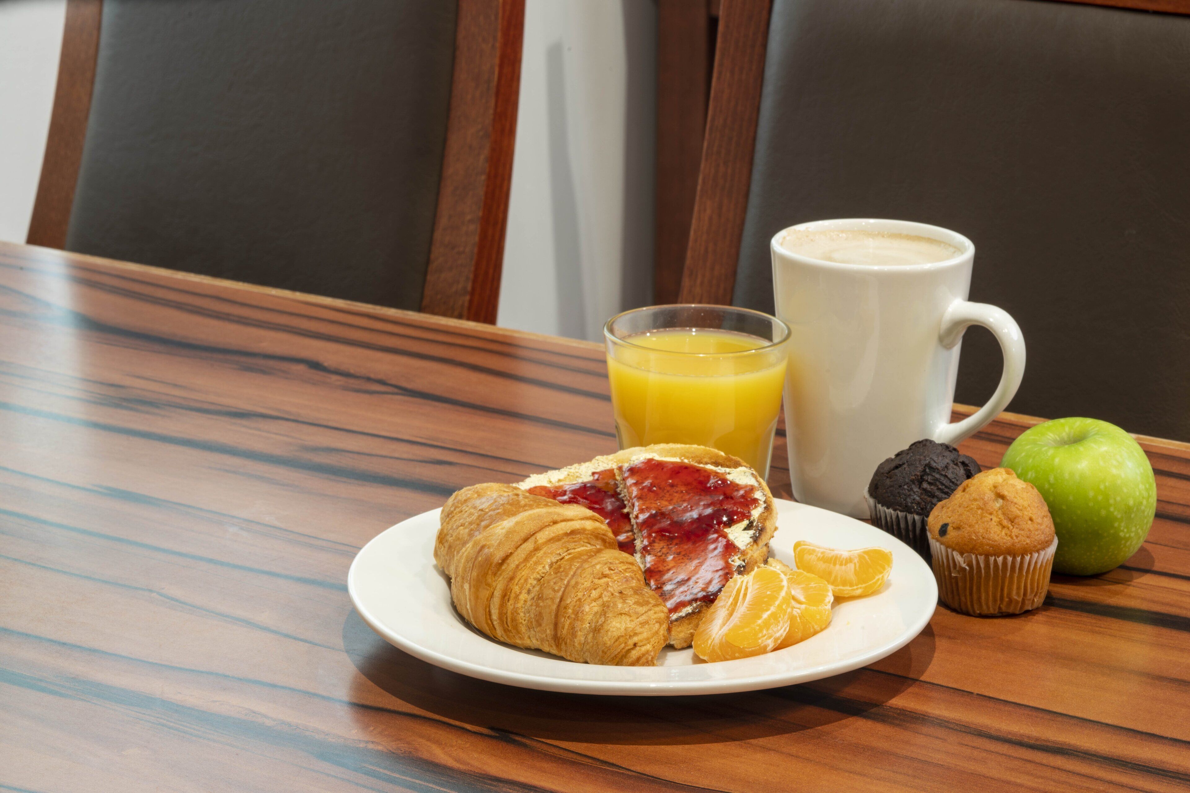 A breakfast setting with croissants, muffins, an apple, a glass of orange juice, and a coffee cup on a wooden table.