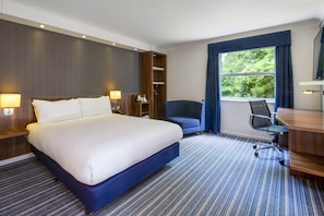 A hotel room with a large bed, a desk with a chair, a wardrobe, and a window with curtains.