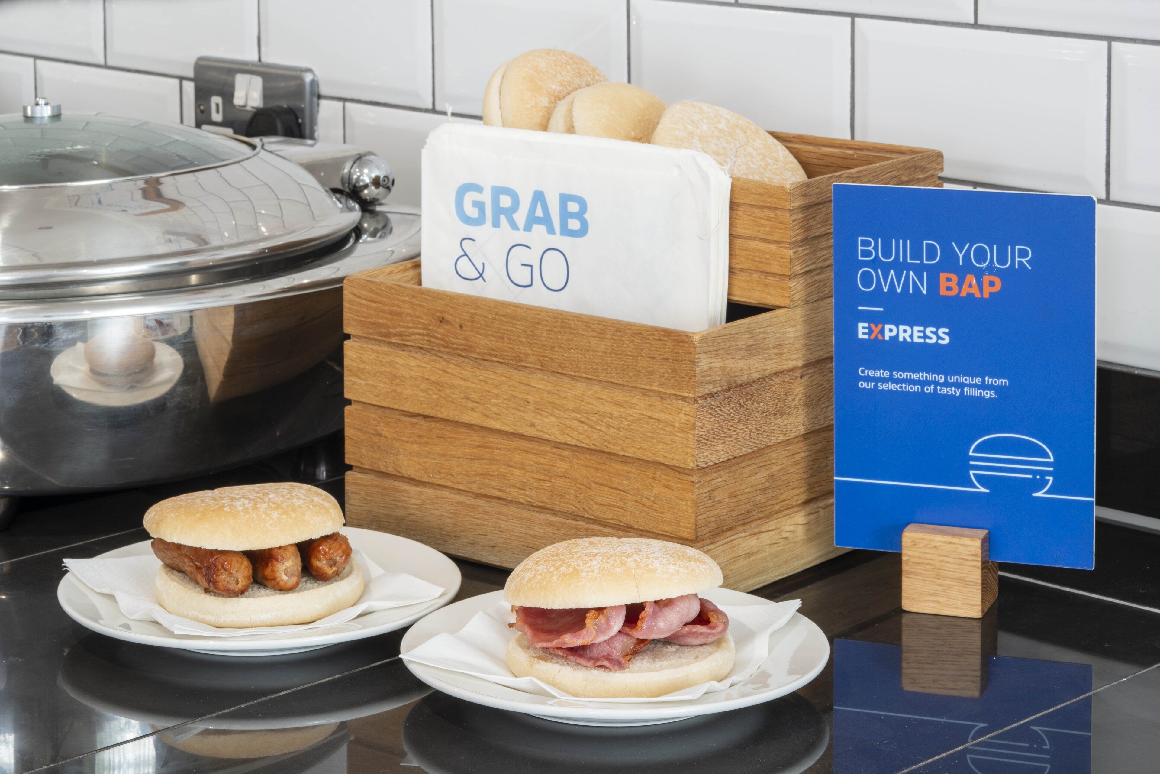 A kitchen counter with a sign that reads "GRAB & GO" and another sign that says "BUILD YOUR OWN BAP EXPRESS." There are two sandwiches on plates in front of the counter.