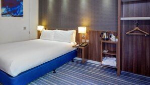 A hotel room with a large bed, two bedside lamps, a wooden headboard, a nightstand with a phone and a kettle, a shelf with towels, and a blue-striped carpet.
