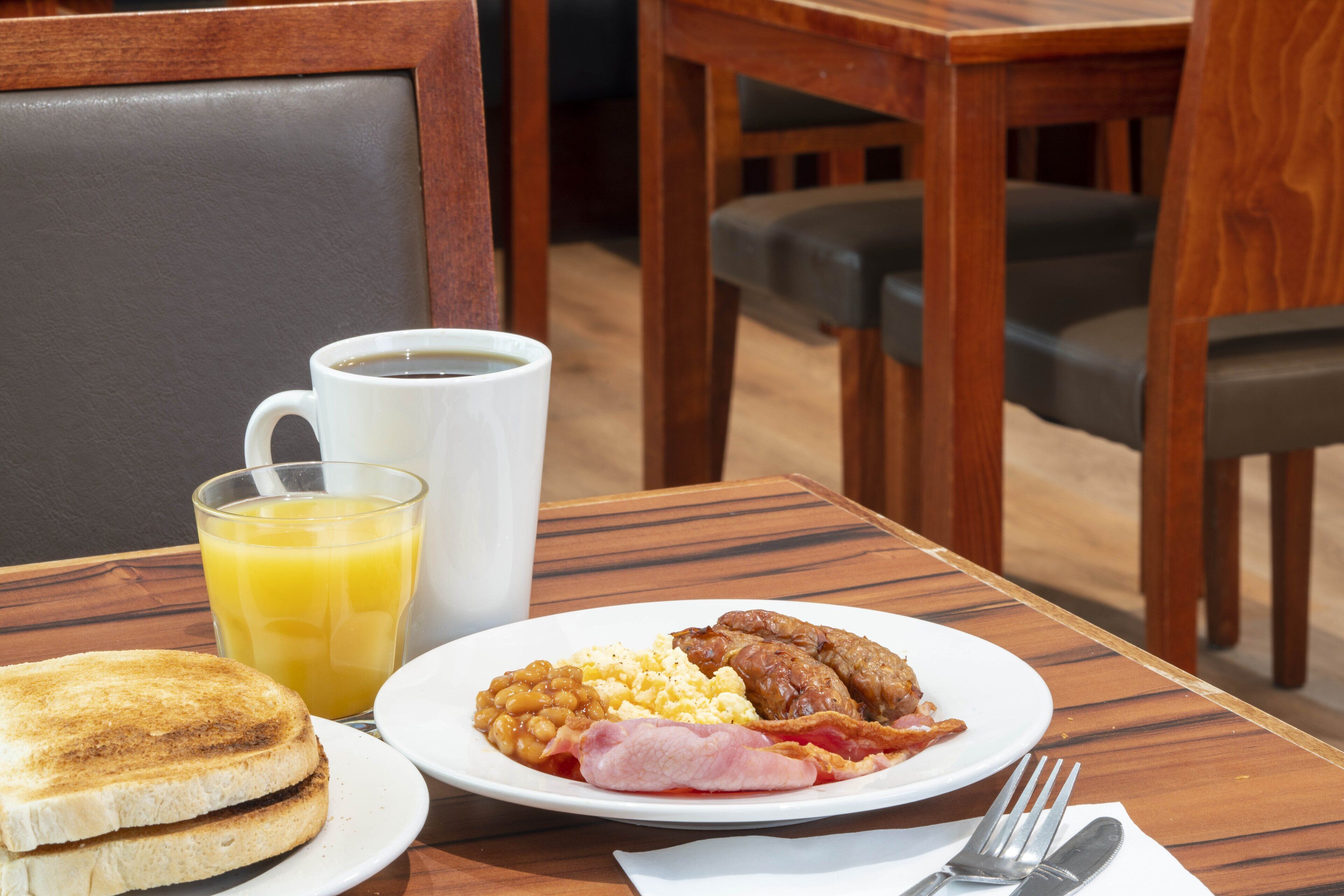 A breakfast setting with toast, eggs, beans, bacon, and a glass of orange juice on a wooden table.