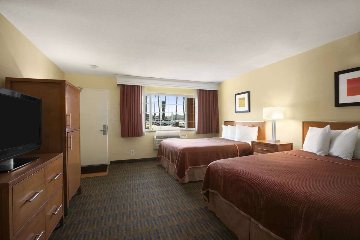 A hotel room with two beds, a TV, a desk, a window with curtains, and a view of the outside.