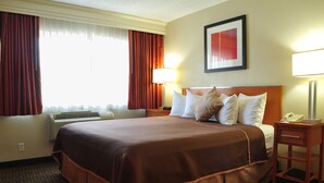 Una habitación de hotel con una cama grande, una mesita de noche con lámpara, un cuadro enmarcado en la pared y una ventana con cortinas.