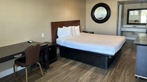 Una camera d'hotel con un letto, una scrivania, una sedia, uno specchio tondo e un bagno con lavandino e specchio.