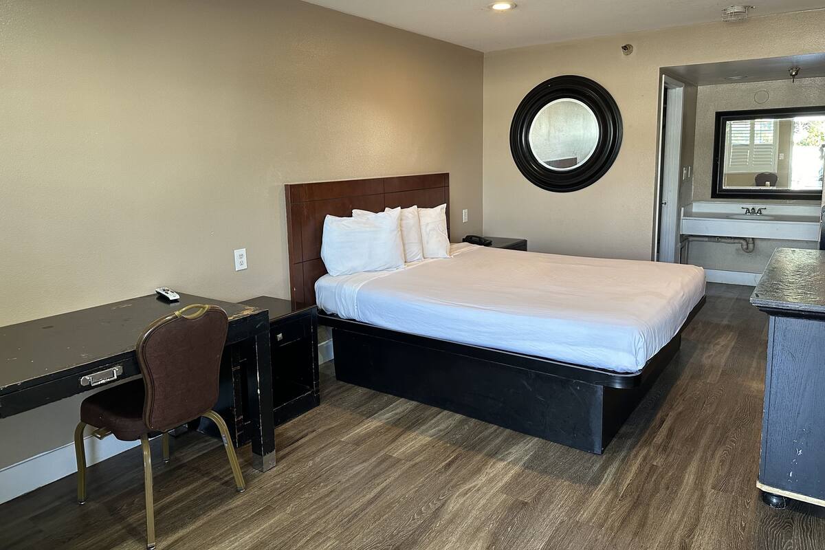 Habitación de hotel con cama, escritorio, silla, espejo redondo y baño con lavamanos y espejo.