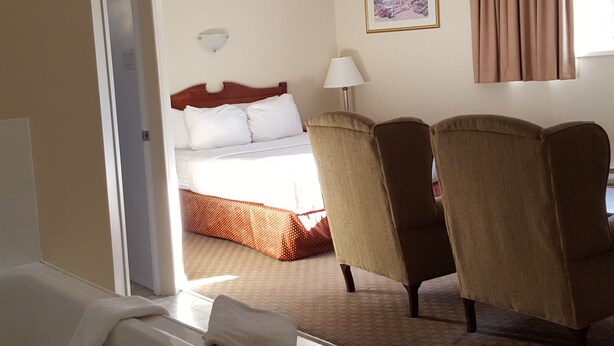 A hotel room with a bed, two armchairs, a desk, a window with curtains, and a painting on the wall. - Knights Inn Lethbridge (Lethbridge)