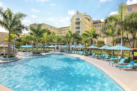 A swimming pool with lounge chairs and umbrellas, surrounded by palm trees and a hotel building.