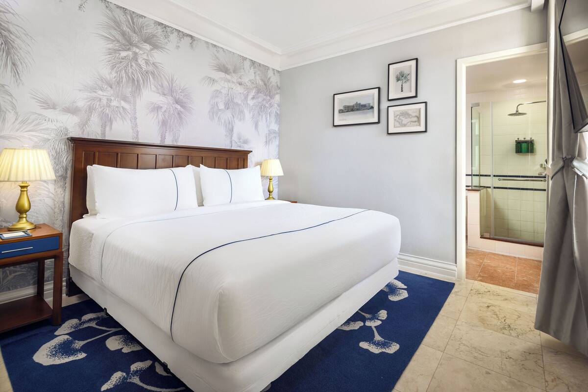 Quarto de hotel com uma cama grande, duas mesas de cabeceira com luminárias, um tapete azul estampado e uma área de chuveiro visível através de uma porta aberta.