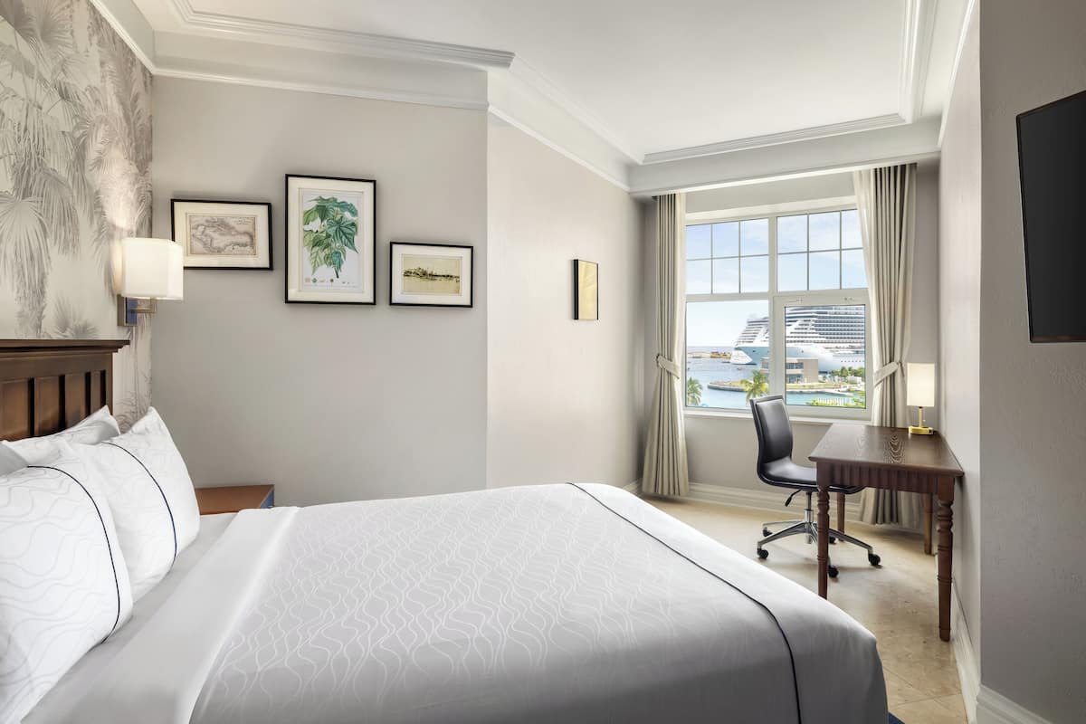 Quarto de hotel com uma cama grande, uma escrivaninha e vista para um navio de cruzeiro pela janela.