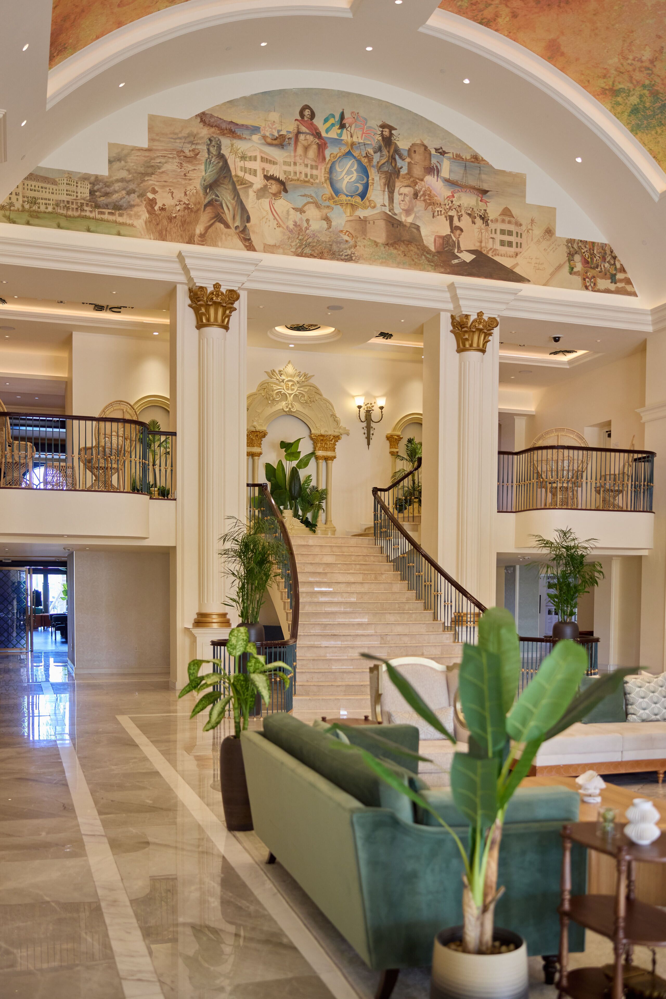 A grand lobby with a marble staircase, a mural on the ceiling, and a green sofa.