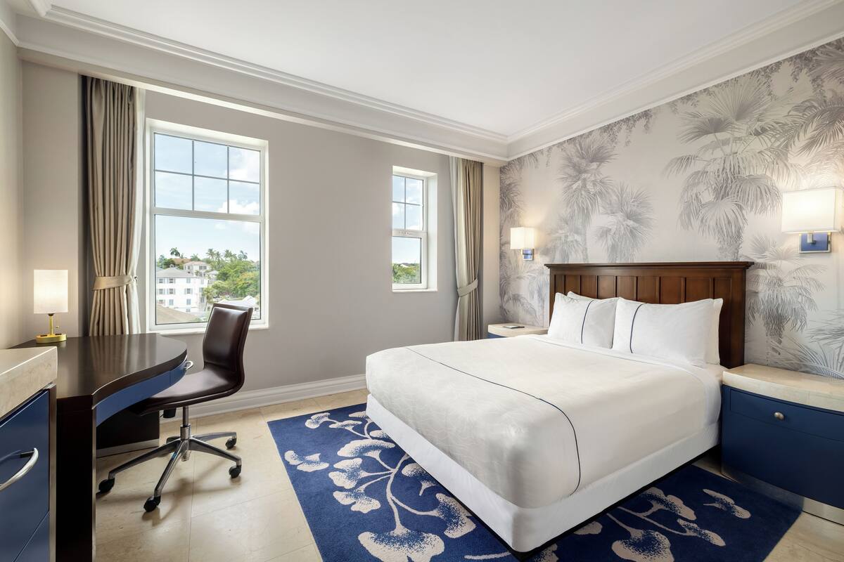 Quarto de hotel com uma cama grande, uma escrivaninha e uma cadeira. Há duas janelas com cortinas, uma parede com um mural de palmeiras e uma mesa de cabeceira com uma luminária.