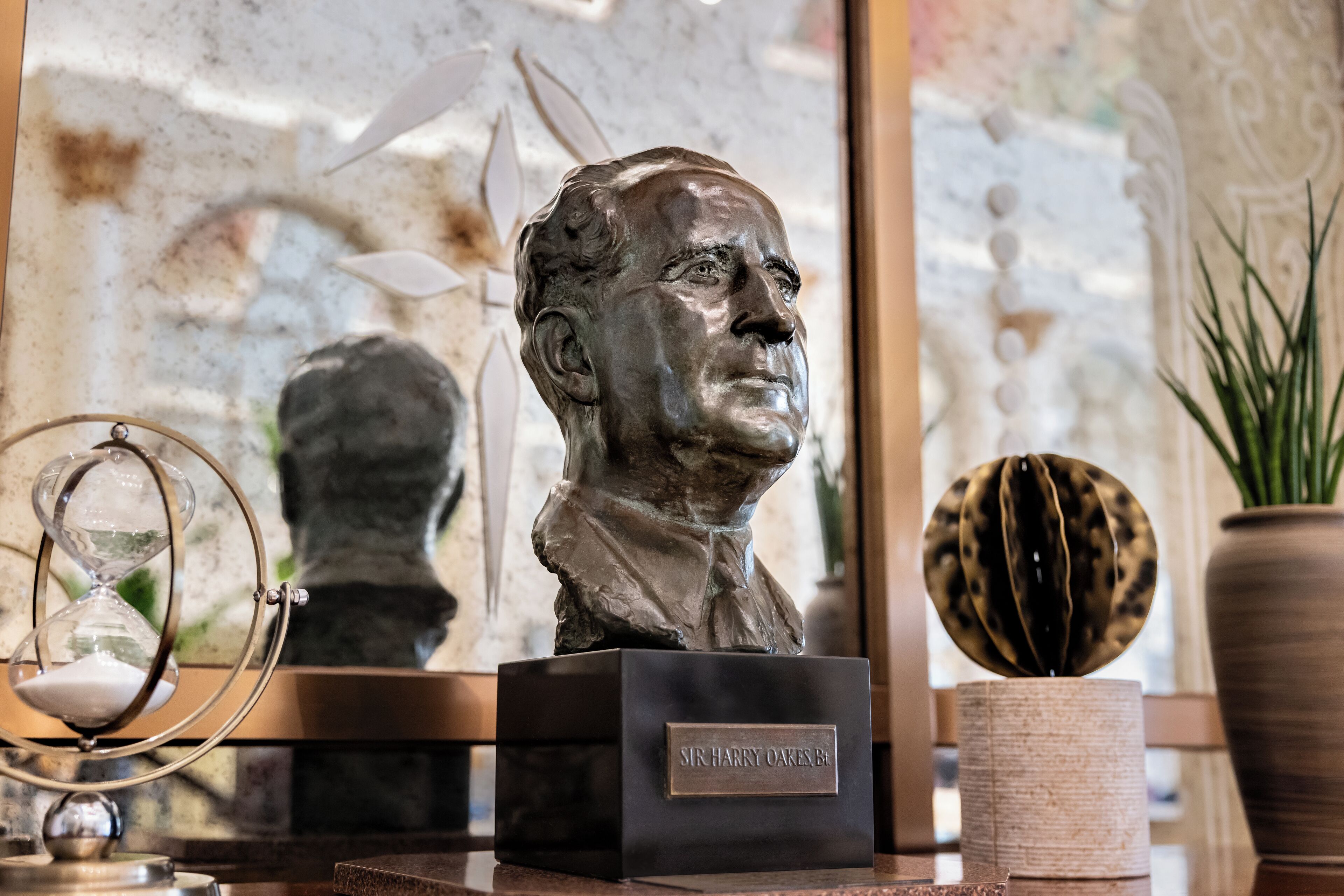 Un busto escultórico sobre un pedestal con una placa, una ampulleta de cristal y una esfera decorativa.
