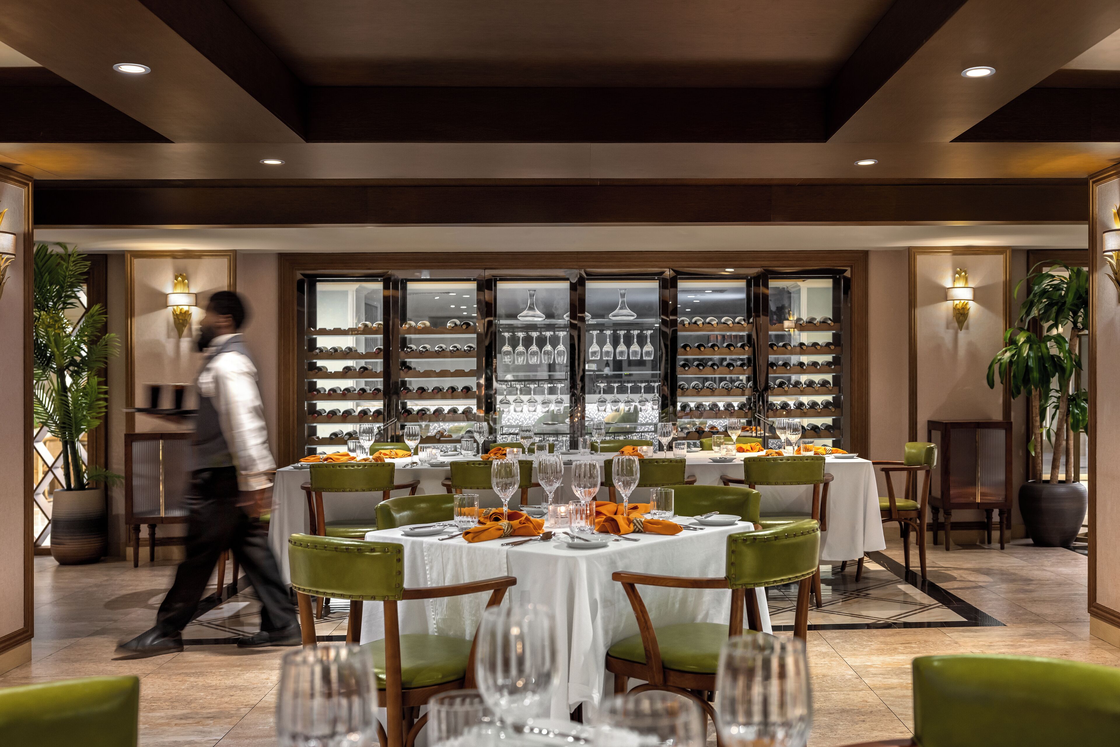 Un restaurante con mesas dispuestas para comer, una exhibición de vinos y un camarero caminando.