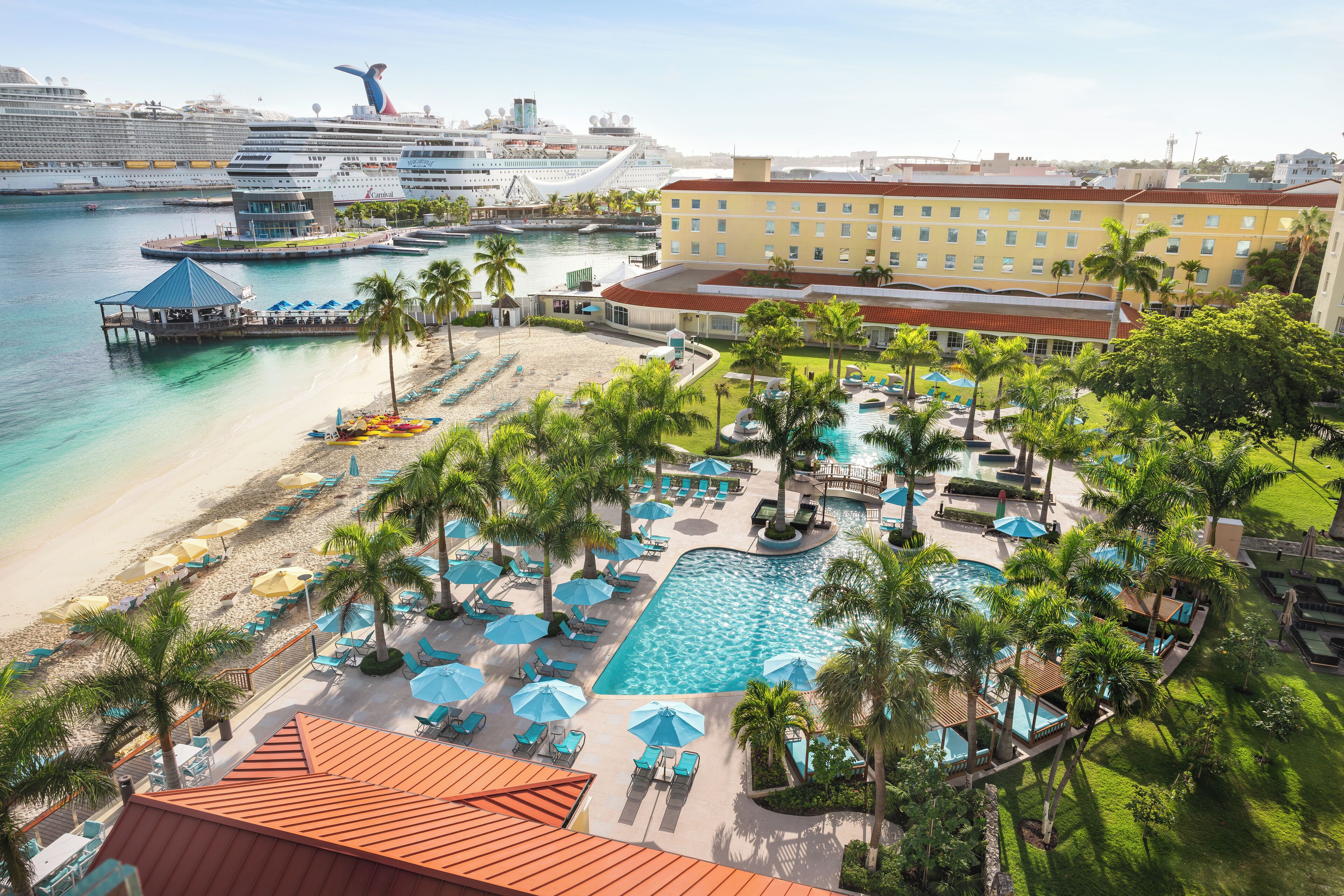 A resort with a pool area, beach, and cruise ships in the background.