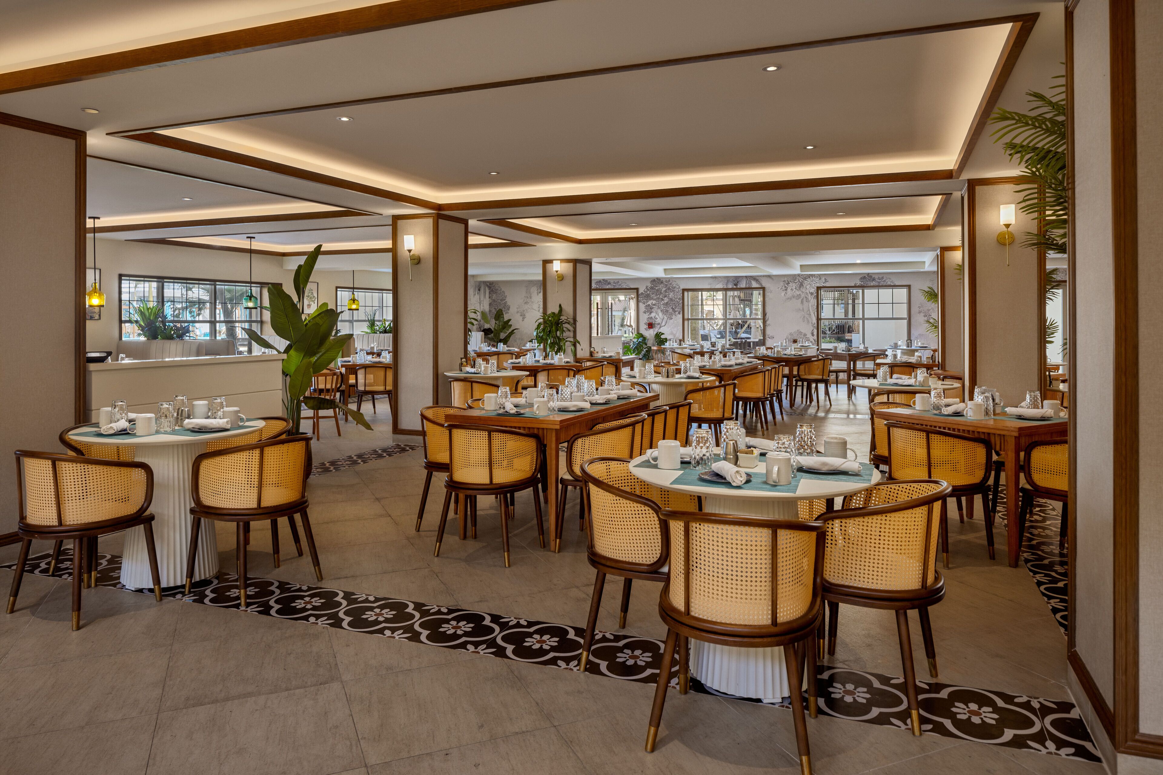 A spacious dining area with multiple round tables and wicker chairs, featuring a patterned floor and large windows.