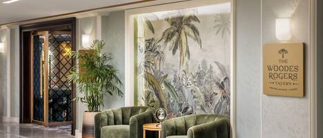 A lobby with a reception desk, two green armchairs, a large wall mural, and a sign for 'The Woodes Rogers Tavern'.