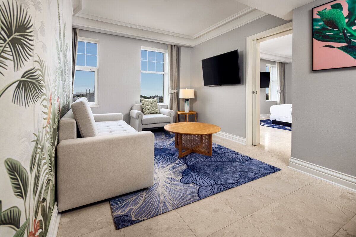 Uma sala de estar moderna com sofá, poltrona e mesa redonda, uma TV de tela plana na parede, e vista da cidade pela janela.