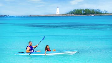 Two people kayaking on a clear blue sea with a lighthouse in the distance.