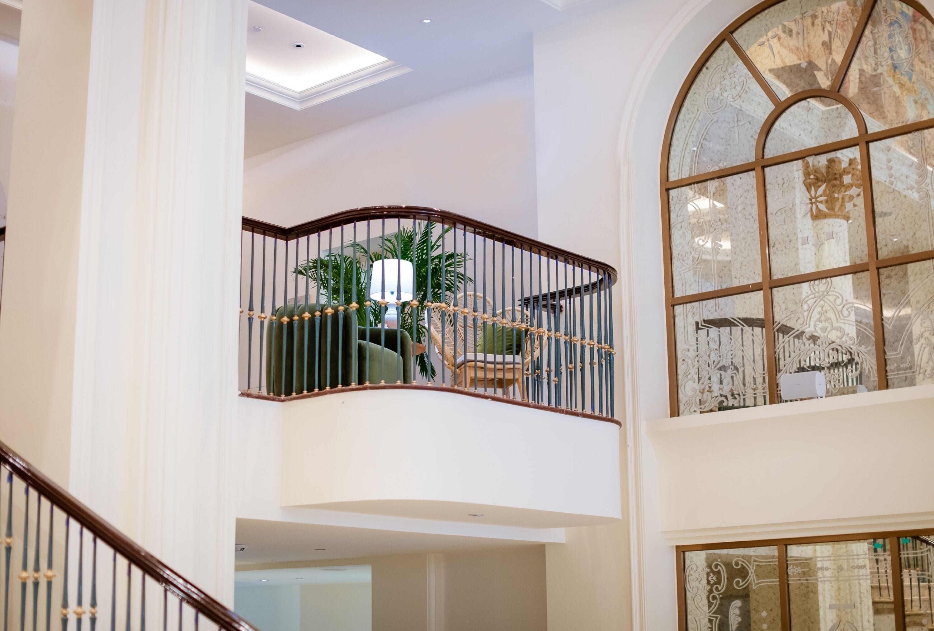 Un interior de varios niveles con una escalera curva, un balcón con plantas y un amplio ventanal de arco con vidrieras decorativas.