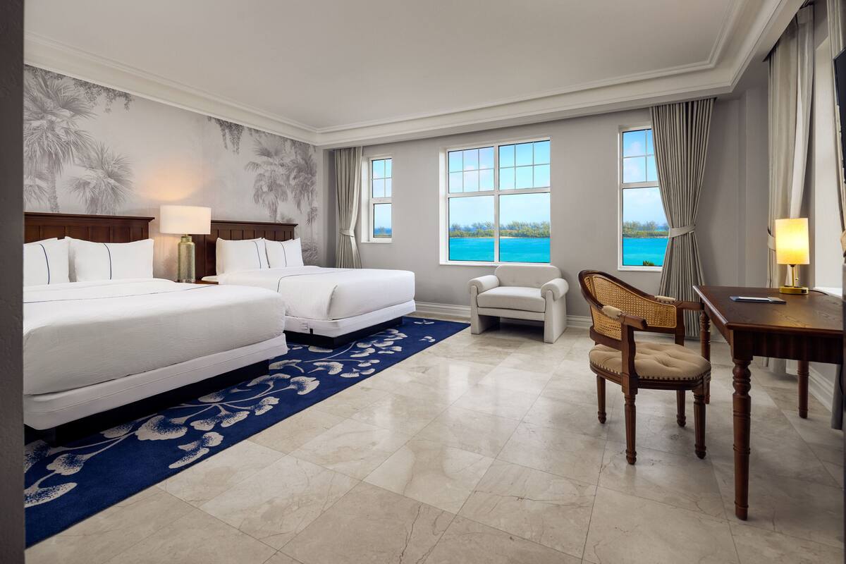 Quarto de hotel com duas camas, uma escrivaninha, uma cadeira e vista para o mar.