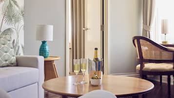 A room with a wooden table, two glasses of white wine, a bottle of champagne, and a bowl of olives. There is a sofa and a wicker chair with cushions.