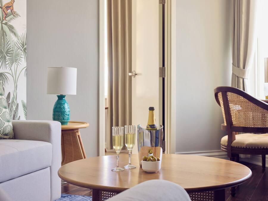 A room with a wooden table, two glasses of white wine, a bottle of champagne, and a bowl of olives. There is a sofa and a wicker chair with cushions.