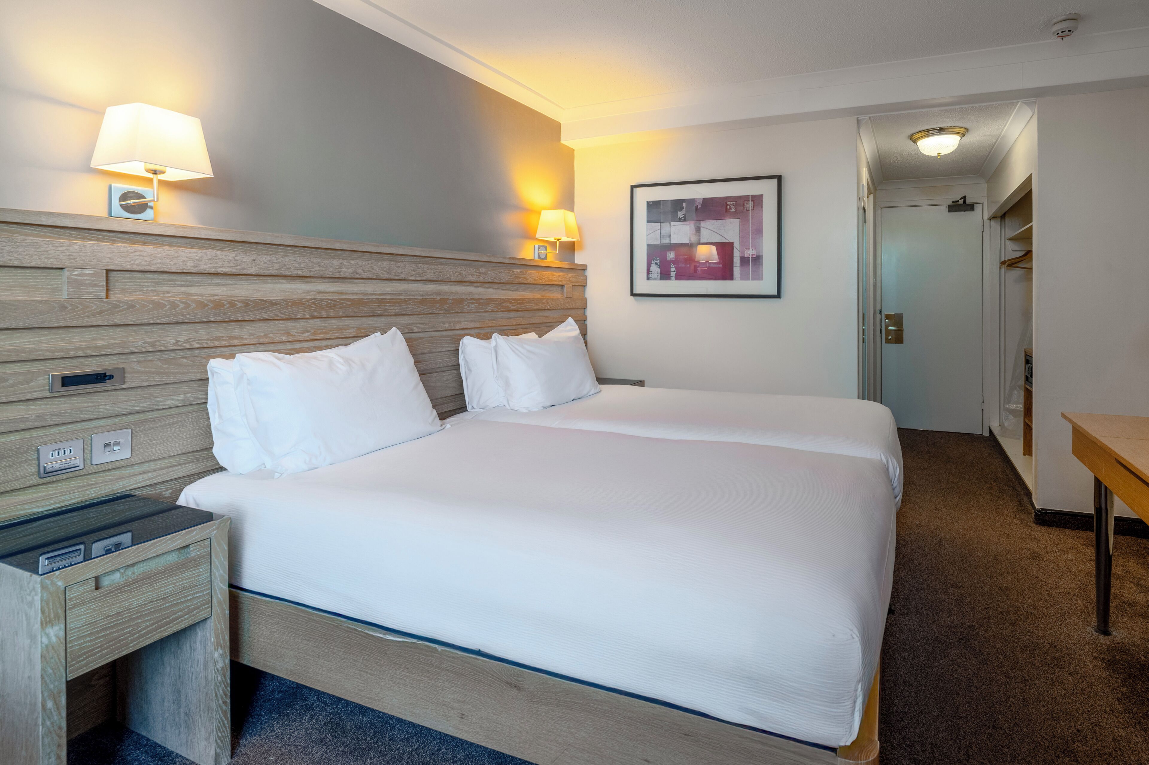 A hotel room with a double bed, bedside lamps, a nightstand, a framed picture on the wall, and a door leading to a closet.
