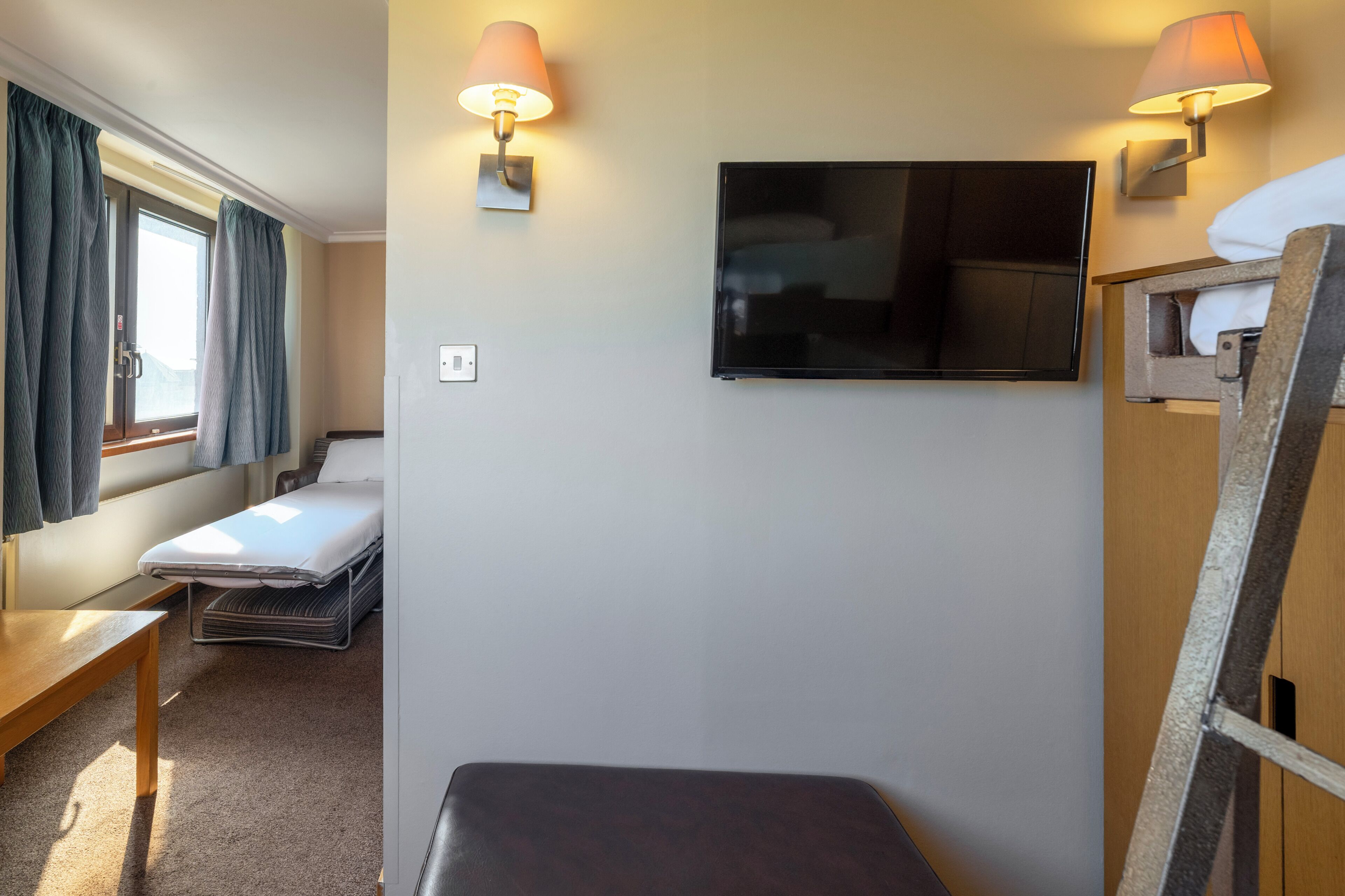 A compact hotel room with a bed, a small table, a bench, a TV mounted on the wall, and two wall-mounted lamps.