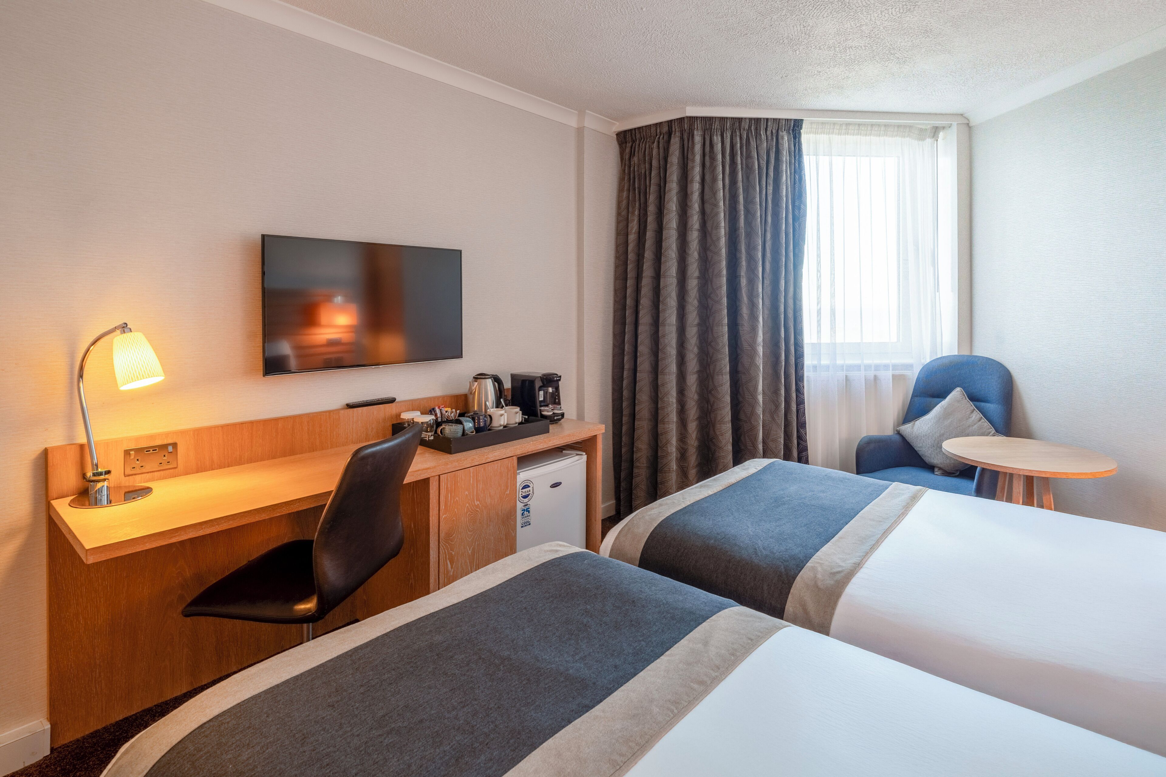 A hotel room with a desk, two beds, a TV, a coffee maker, and a small table.