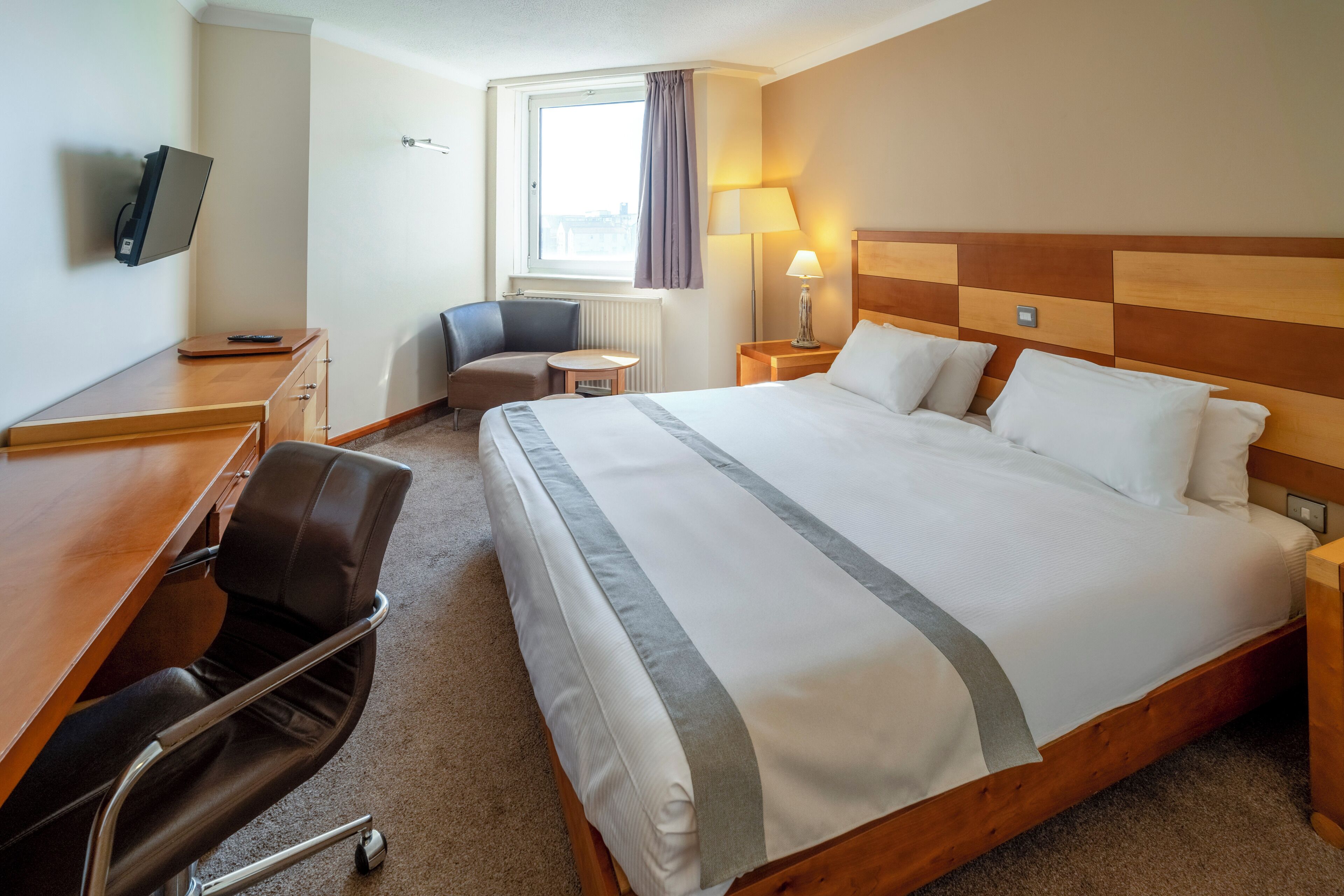 Ein Hotelzimmer mit einem großen Bett, einem Schreibtisch mit Stuhl, einem Fernseher, einer Lampe und einem Fenster mit Vorhängen.