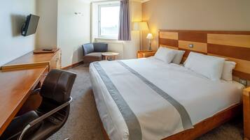 A hotel room with a large bed, a desk with a chair, a TV, a lamp, and a window with curtains.