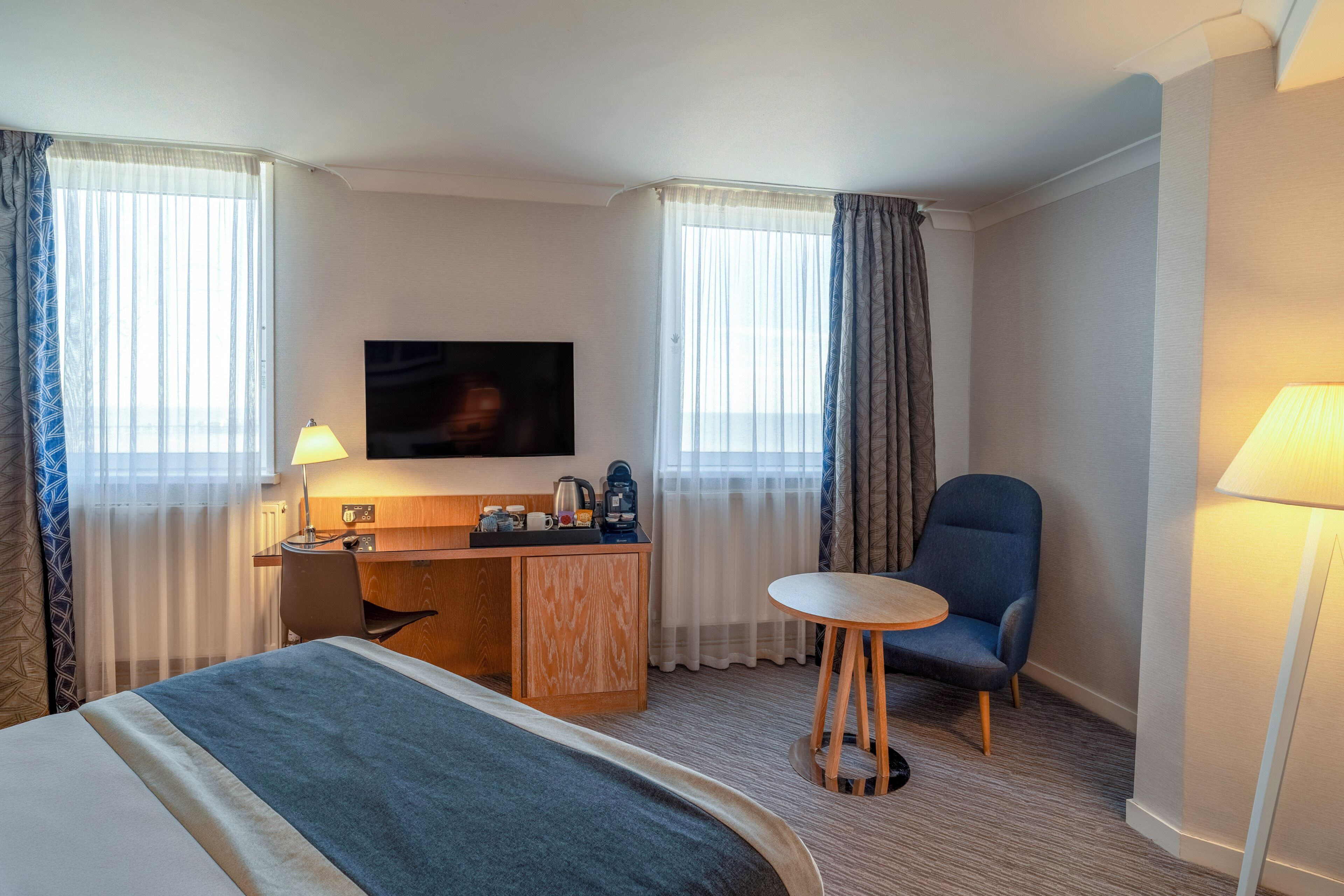 A hotel room with a bed, a desk, a chair, a TV, and a lamp.