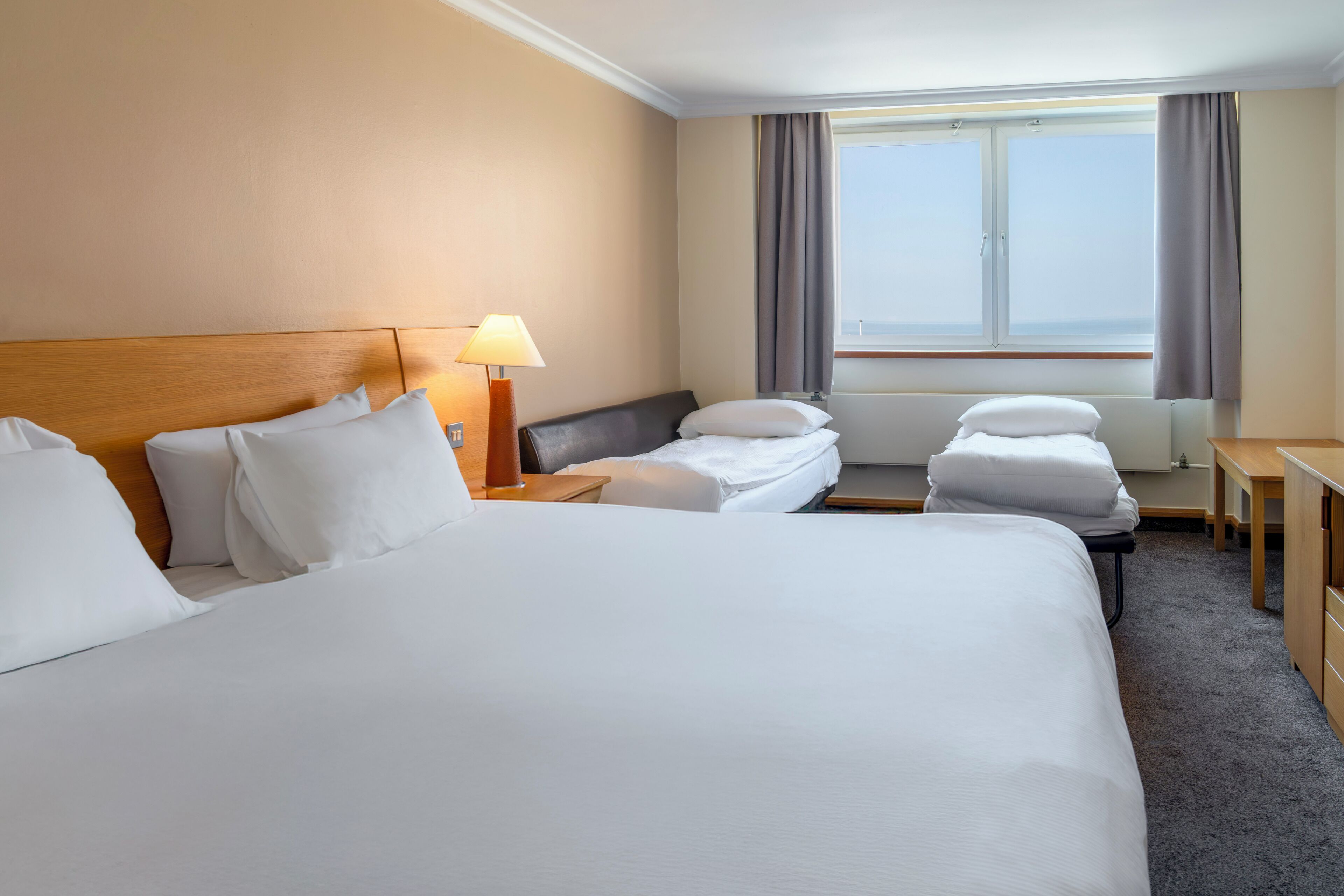 A hotel room with a large bed, two bedside tables, a desk, a lamp, a window with curtains, and a view of the sea.