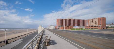 Una carretera costera con un puente peatonal, una playa y un edificio rojo grande al fondo.