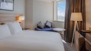 A hotel room with a large bed, a small table, a sofa with cushions, a lamp, and a window with curtains.