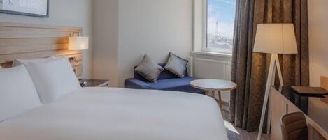 Habitación de hotel con una cama grande, una mesita pequeña, un sofá con cojines, una lámpara y una ventana con cortinas.