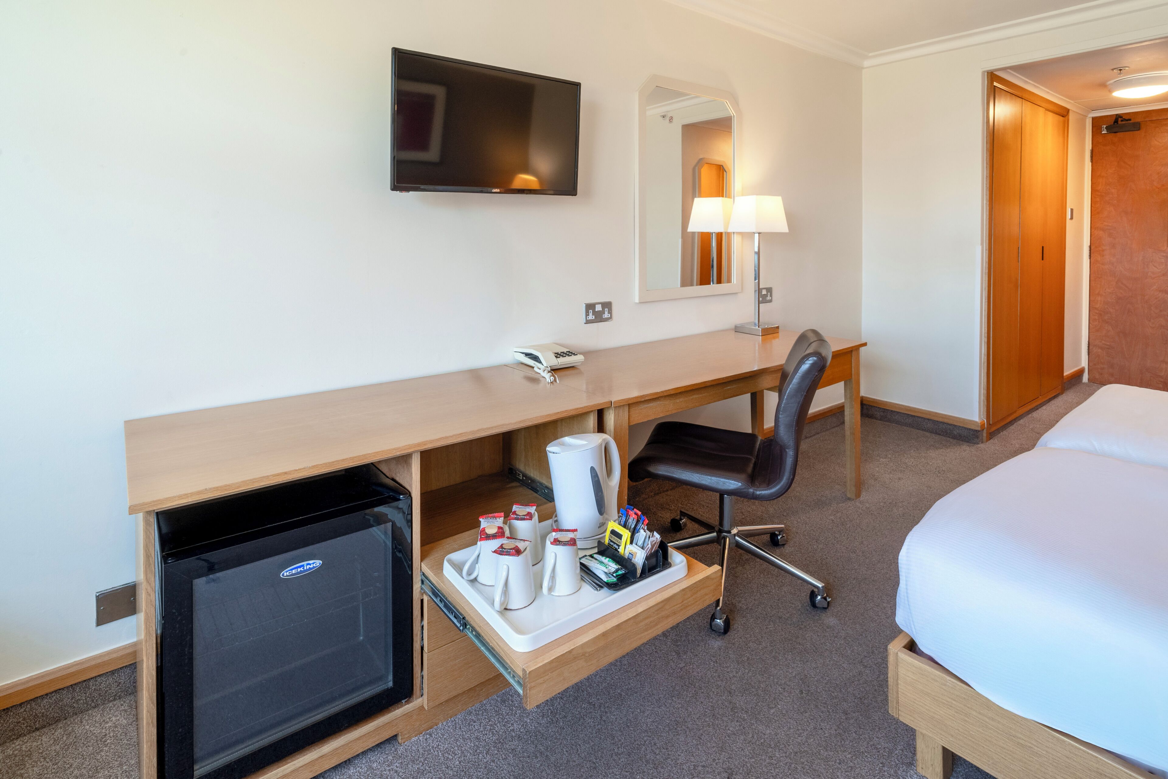 Habitación de hotel con una cama, un escritorio con computadora, televisión, espejo, lámpara y minibar.