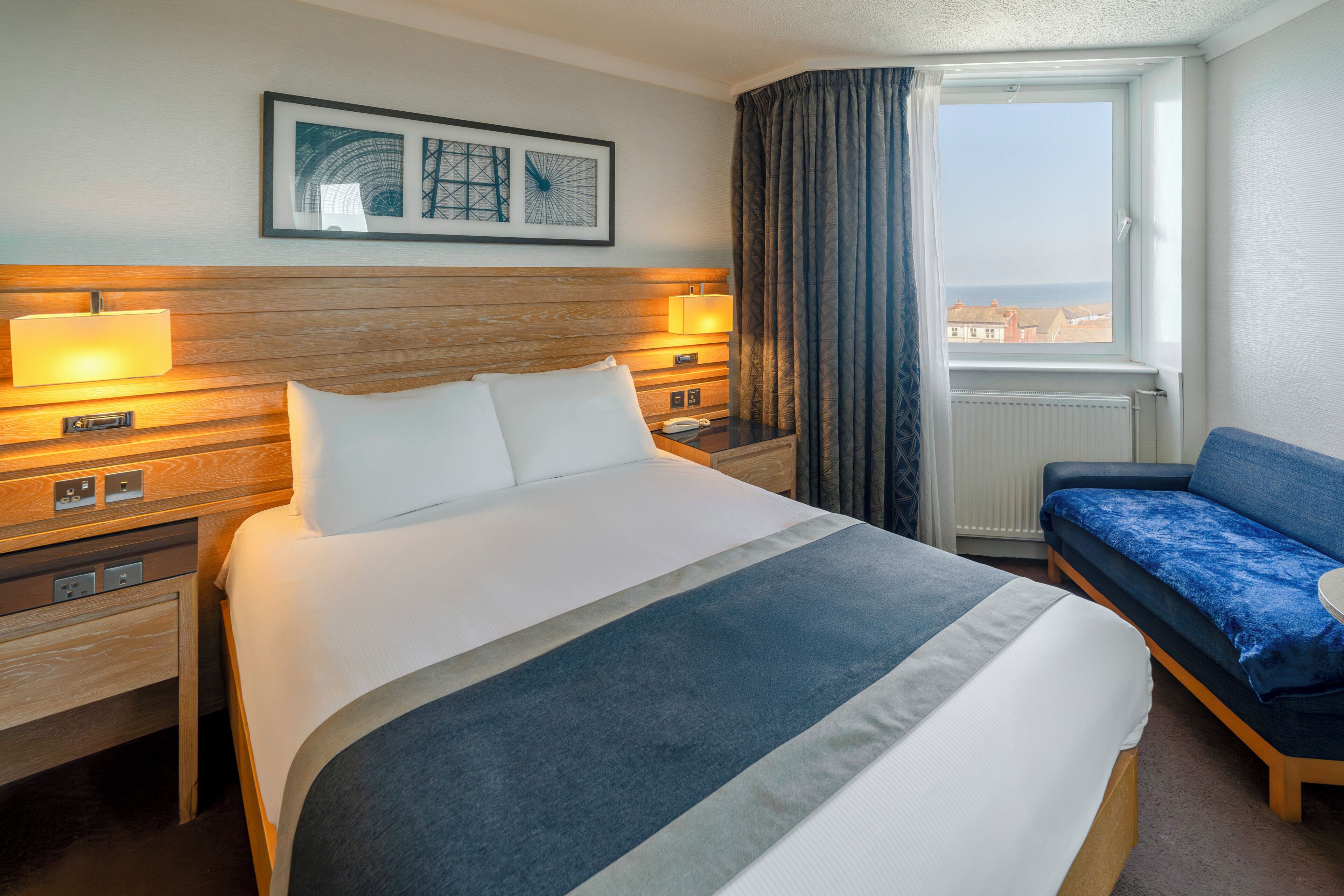 A hotel room with a large bed, bedside tables, a desk, a chair, a window with curtains, and framed artwork on the wall.