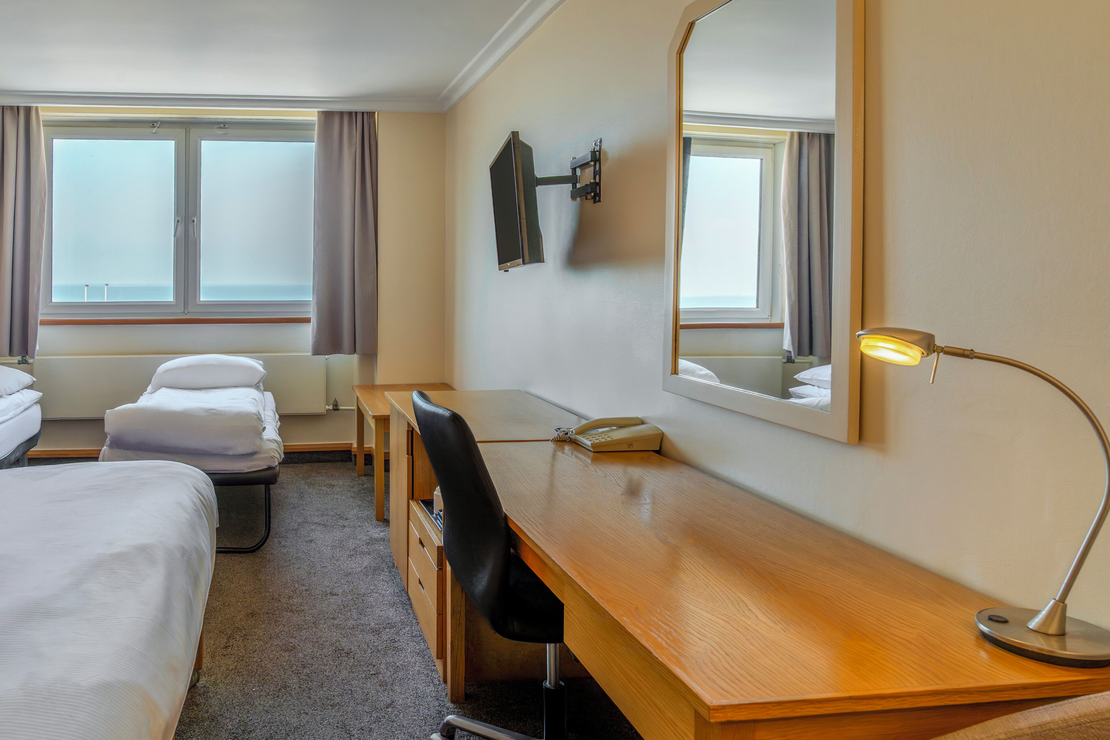 A hotel room with a bed, a desk, a chair, a lamp, a mirror, and a window with curtains.