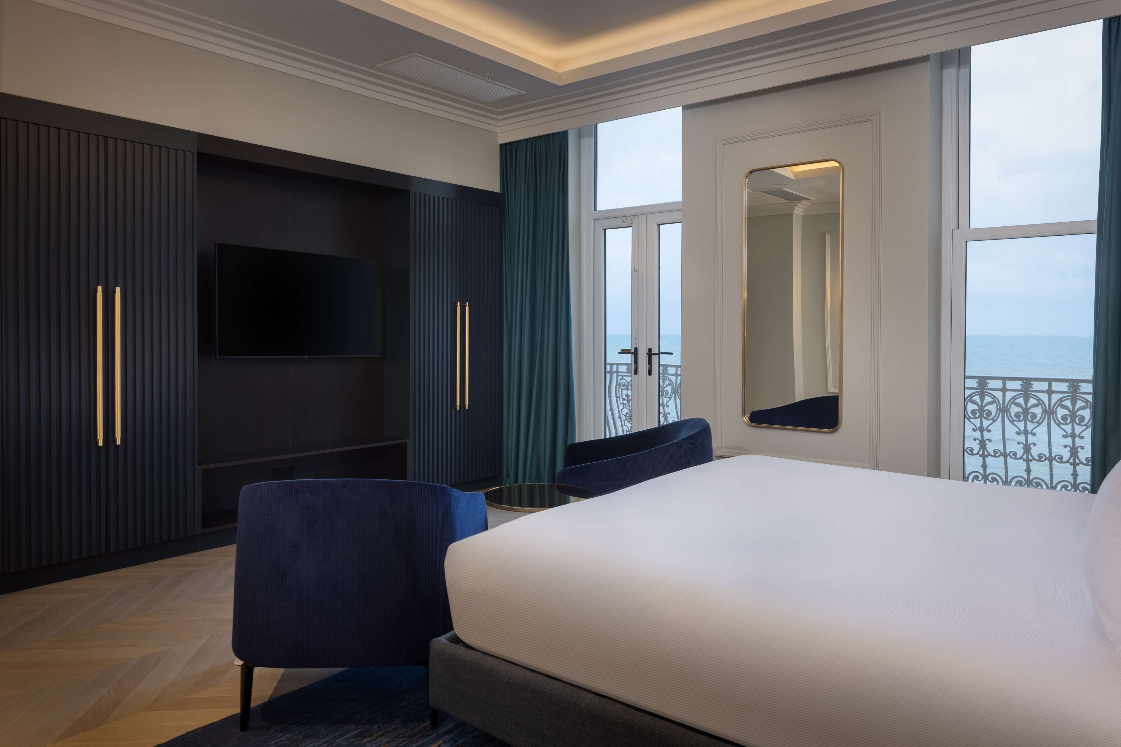 A modern hotel room with a large bed, a flat-screen TV, dark wood paneling, and a balcony with a view.